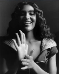 Black and white portrait of Olivia Dean smiling with hands raised, wearing a zip front top with a knit collar against a dark background.