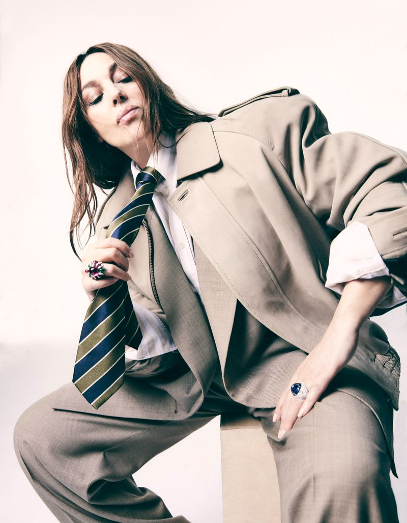 A woman in a beige suit and striped tie poses, seated on a neutral box in a fashion-style portrait shot in a photo studio with soft lighting.