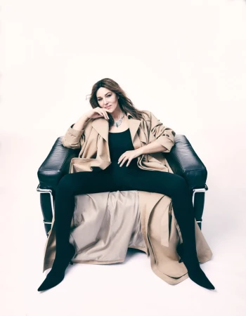 Monica Bellucci sits in a black leather chair wearing a black top and pants, a beige trench coat, and jewelry against a white backdrop.