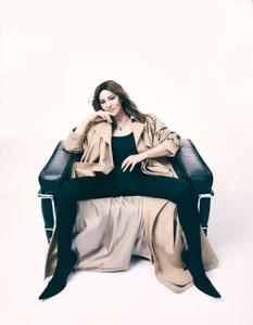 Monica Bellucci seated in a black leather armchair wearing a beige trench coat and black catsuit