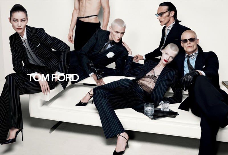 Group of fashion models in tailored dark pinstripe suits and sunglasses posing on and around a white modern bench.