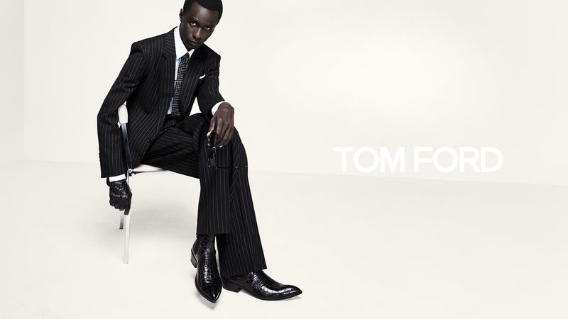 Model Malick Bodian seated in a black pinstripe suit and textured leather boots against a white studio background for Tom Ford.