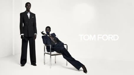 Two models wearing sharp tailored dark suits and gloves posing in a minimalist white studio for Tom Ford.
