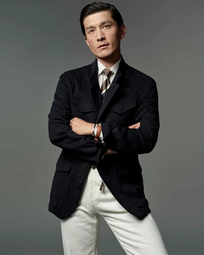 Model Daisuke Ueda in a black tailored field jacket, tie, and white trousers with arms crossed against a grey studio backdrop.