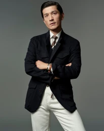 Model Daisuke Ueda in a black tailored field jacket, tie, and white trousers with arms crossed against a grey studio backdrop.