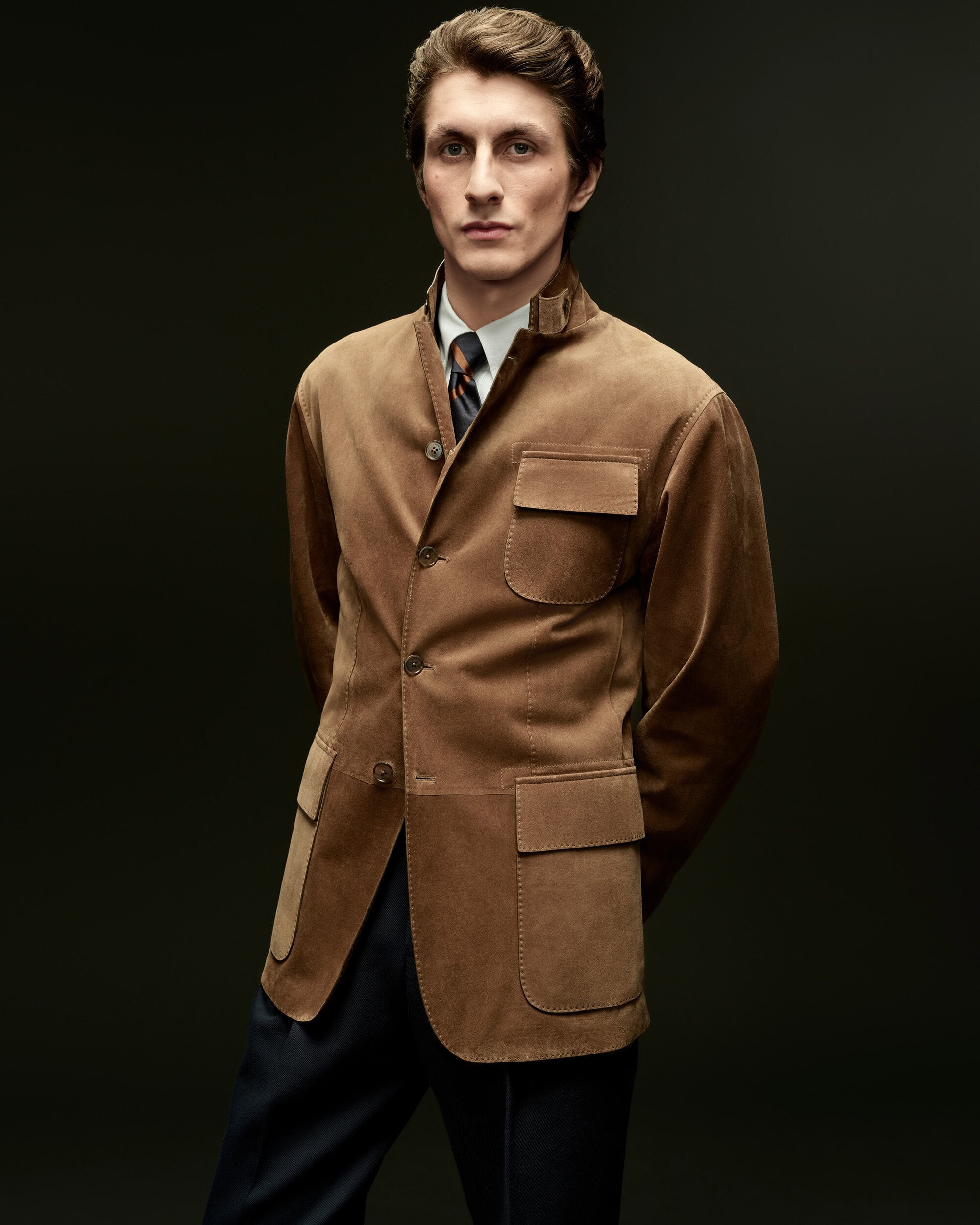 Model Henry Kitcher posing in a tan suede safari jacket, white shirt, and tie against a dark studio background.