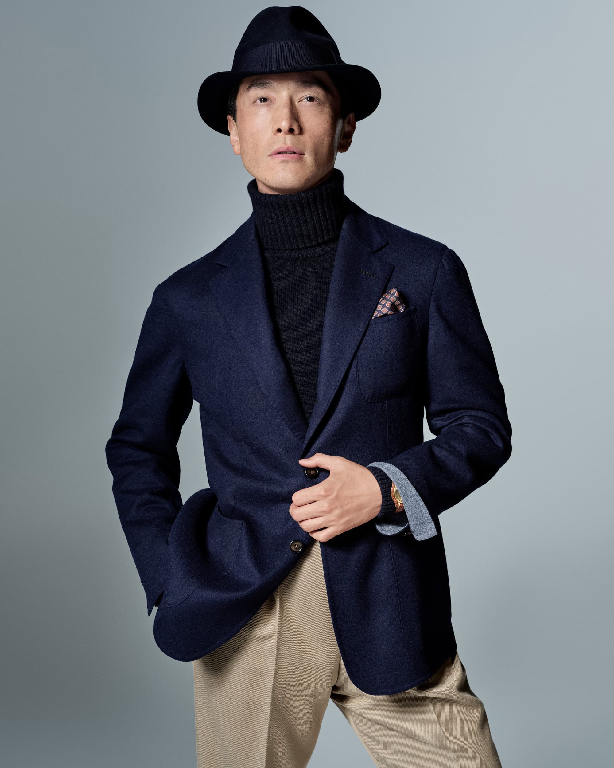 Model Daisuke Ueda wearing a navy blazer, black turtleneck, beige trousers, and a fedora against a studio backdrop.