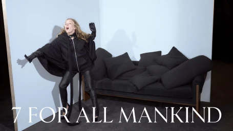Chloë Sevigny poses in an all-black hooded cape and leather pants next to a sofa filled with cushions against a pale blue set.
