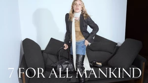 Chloe Sevigny posing on a dark grey sofa wearing a blazer, denim jeans, long white scarf, and thigh-high black leather boots.