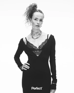 Model Natasha Poly wearing a black plunging dress, lace bralette, and chain necklaces against a white studio background.