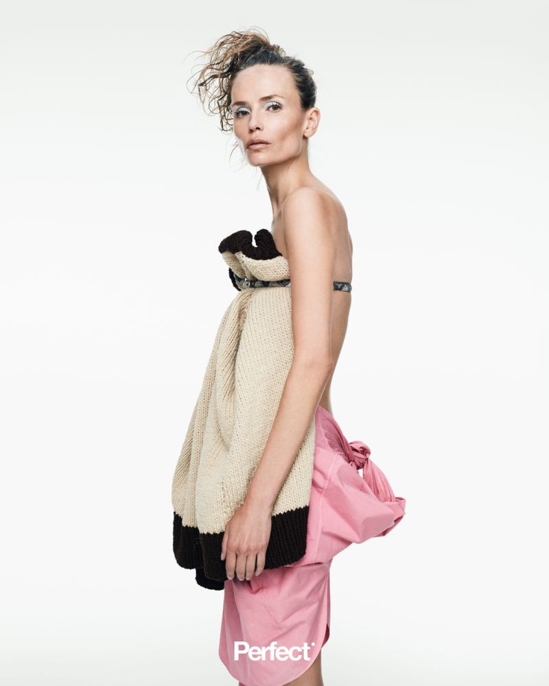 Model Natasha Poly posing in profile wearing a cream knit top and pink draped skirt against a white studio background.