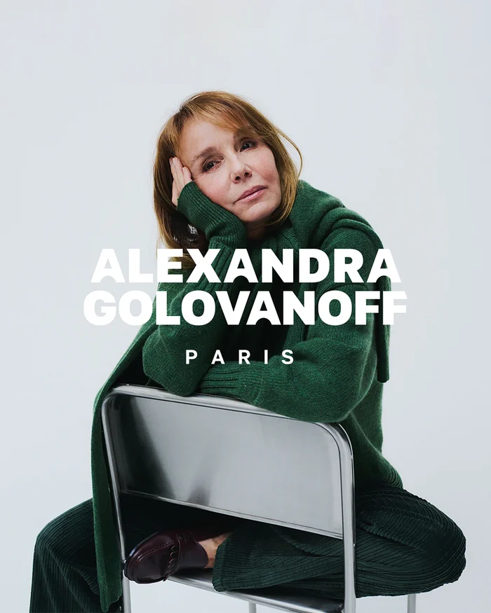 Philippine Leroy-Beaulieu seated backwards on a metal chair wearing a green knit sweater against a seamless white studio backdrop.