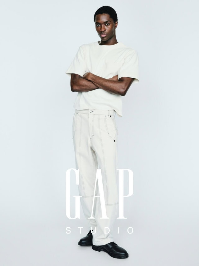Alton Mason posing with arms crossed in an off-white t-shirt and wide-leg trousers against a white studio backdrop.