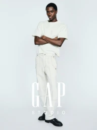 Alton Mason posing with arms crossed in an off-white t-shirt and wide-leg trousers against a white studio backdrop.