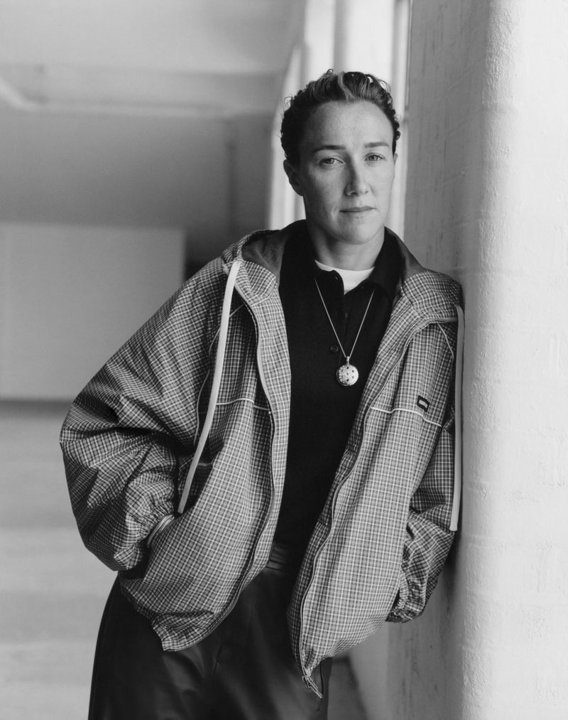 Portrait of a woman with short hair, wearing a patterned jacket and necklace, leaning against a column in a hallway.