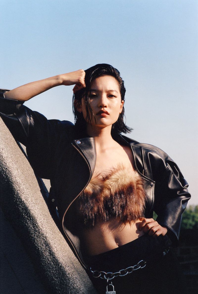 Adult female fashion model in a dark leather jacket, fur top, black pants, and metal chain posing outdoors under a bright blue sky.