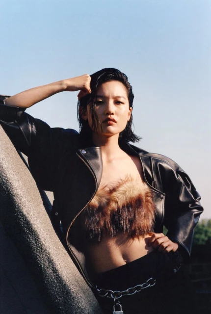 Adult female fashion model in a dark leather jacket, fur top, black pants, and metal chain posing outdoors under a bright blue sky.
