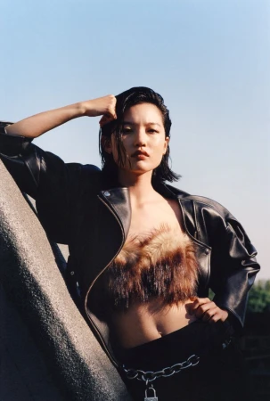 Adult female fashion model in a dark leather jacket, fur top, black pants, and metal chain posing outdoors under a bright blue sky.