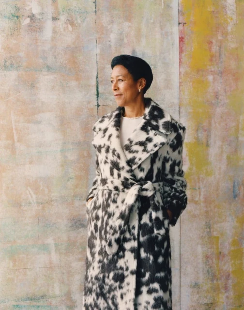 Elegant older woman in a Dalmatian-print coat poses against a colorful abstract background. The image has a stylish, artistic feel.