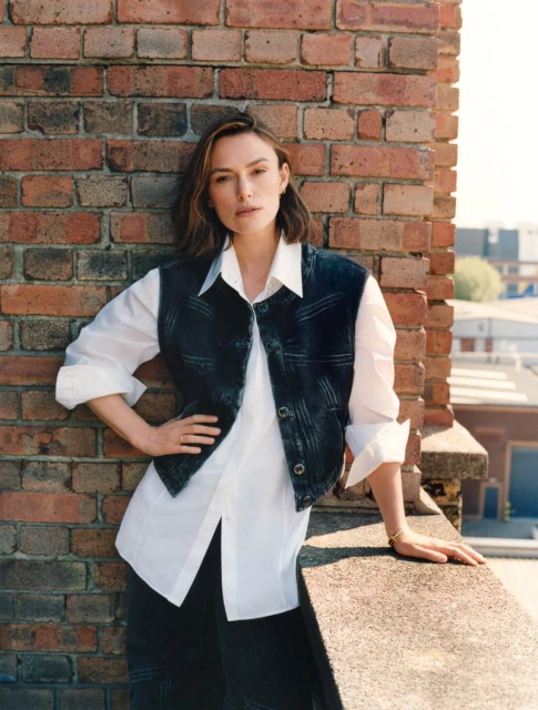 Portrait of an adult woman in a denim vest and white shirt posing against a brick wall outdoors on a sunny day.