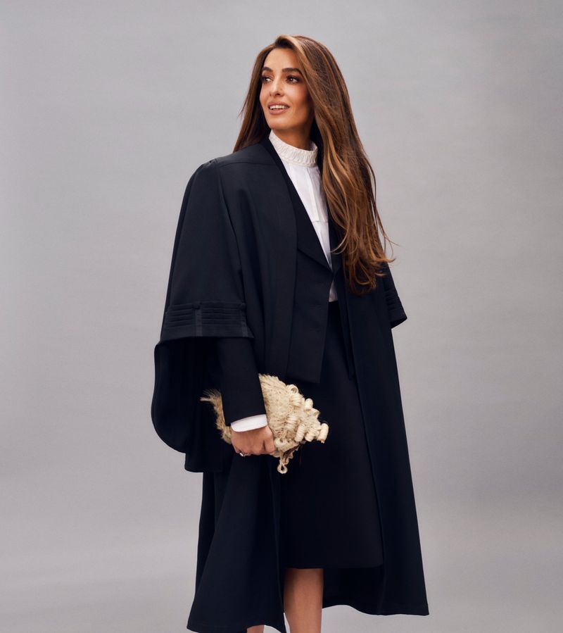 A woman in a black academic gown smiles and looks left, holding a traditional barrister's wig in front of a grey studio backdrop.