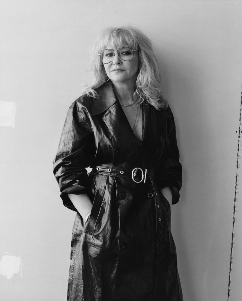 Black and white portrait of a blonde adult woman wearing glasses and a dark trench coat, posing indoors against a light-colored wall.