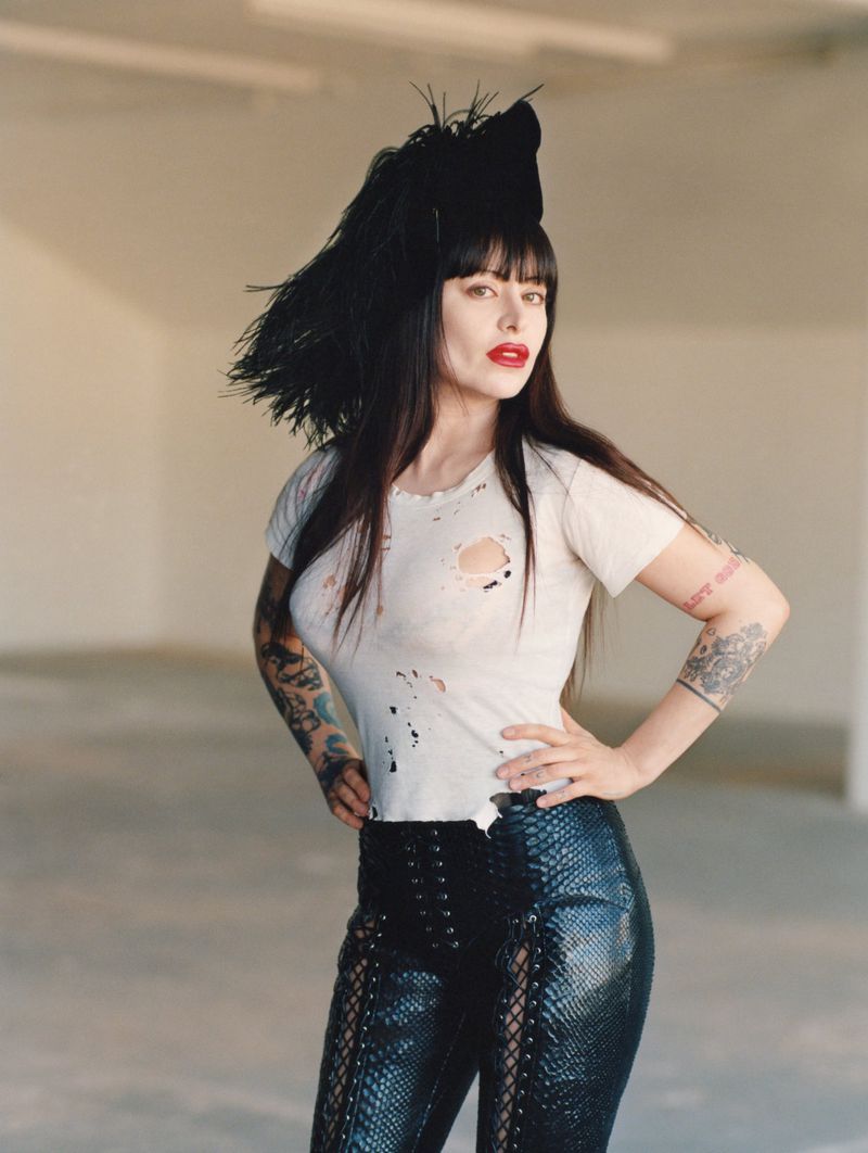 Woman in a white t-shirt and black pants poses in a photo studio with neutral walls, showcasing fashion and edgy style with arm tattoos.