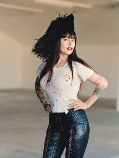 Woman in a white t-shirt and black pants poses in a photo studio with neutral walls, showcasing fashion and edgy style with arm tattoos.