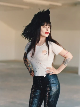 Woman in a white t-shirt and black pants poses in a photo studio with neutral walls, showcasing fashion and edgy style with arm tattoos.