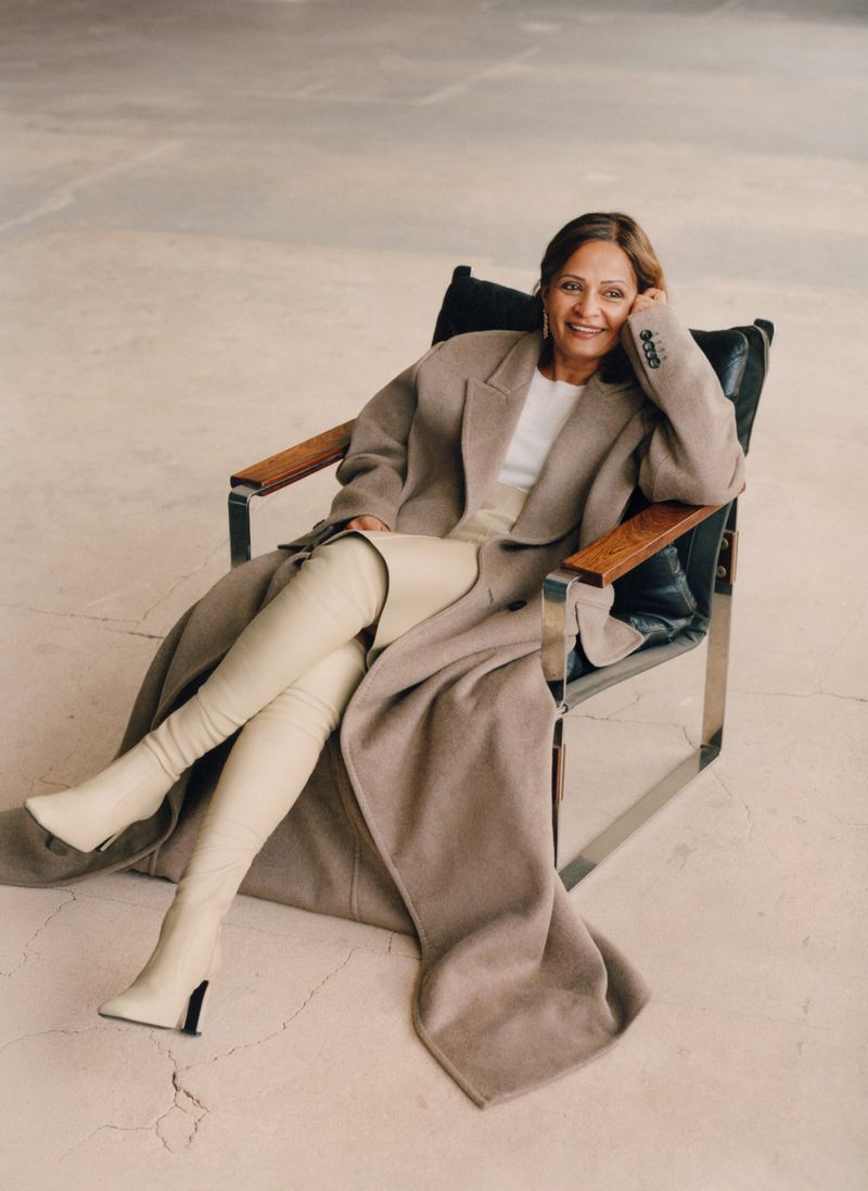 Adult woman sitting in a black chair wearing a long coat and boots in an indoor setting with a neutral-toned background.