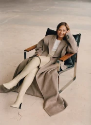 Woman seated in a modern armchair wearing a long taupe coat and tall cream boots against a concrete background.