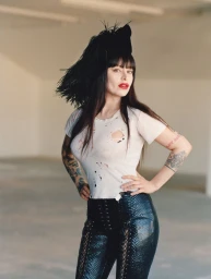 Woman wearing a distressed white t-shirt, black lace-up pants, and a feathered headpiece posing in a bright minimalist interior.