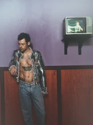 Harry Styles in a metallic embellished jacket and jeans leaning against a purple wall with a mounted television.