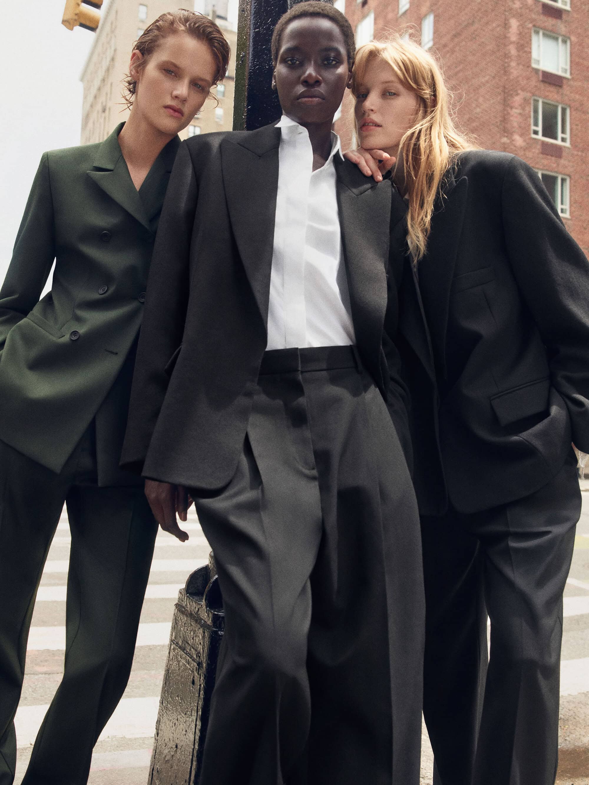 Three models wearing oversized tailored black and dark green suits standing together on an urban city street.