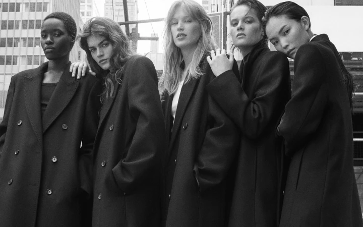 Five female models standing together wearing oversized black tailored coats in a monochrome fashion campaign for COS.