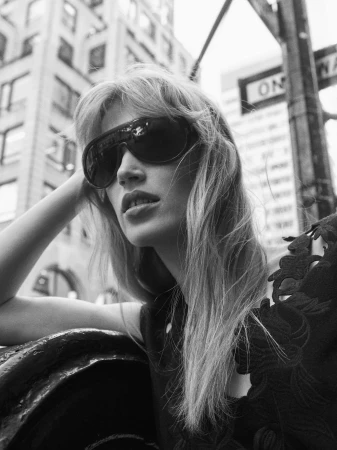 Black and white low-angle portrait of a woman in large shield sunglasses leaning against an urban railing with buildings in the background.