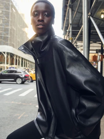 Model Awar Odhiang wearing an oversized black leather jacket with a high collar on an urban street beside scaffolding.