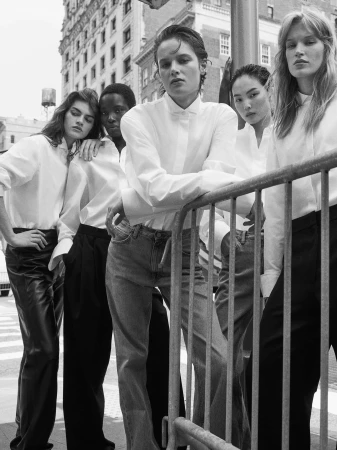 Five models wearing crisp white button-down shirts and tailored trousers stand together beside a metal street barrier in black and white.