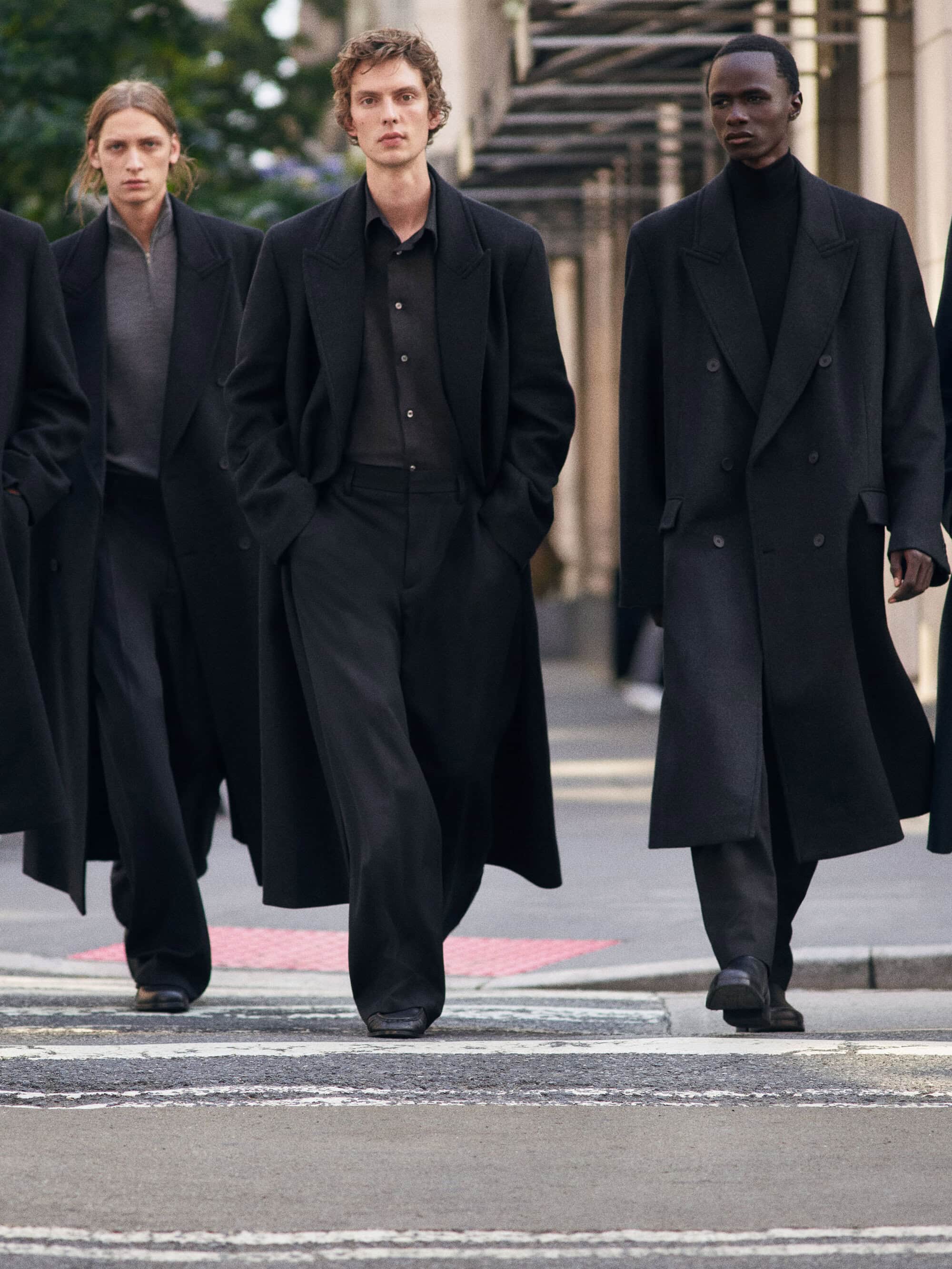 Male models walking across a city street wearing tailored black overcoats and trousers for a fashion campaign.