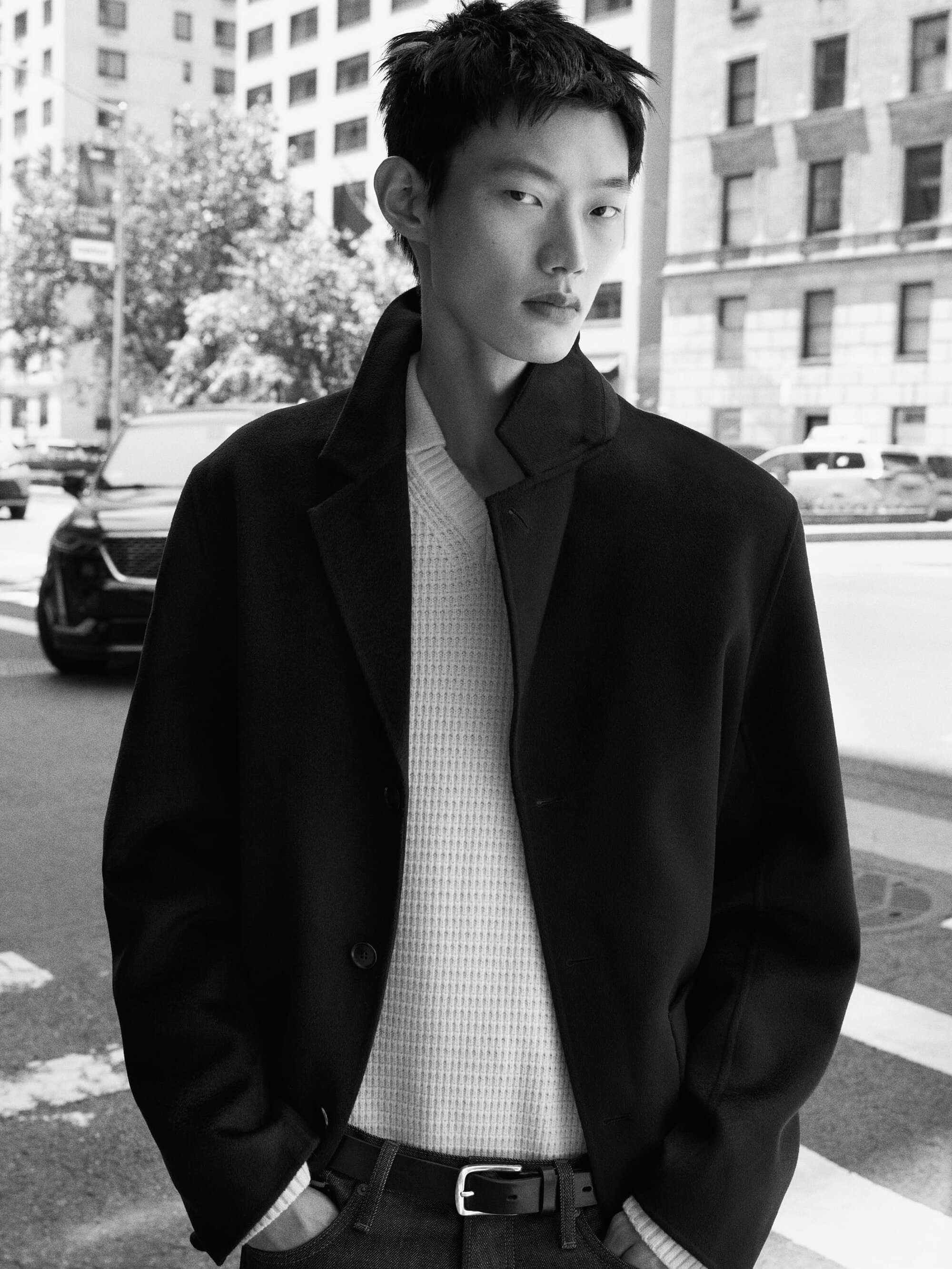 East Asian male model posing on a city street wearing a black coat over a white textured knit sweater for COS Fall 2026.