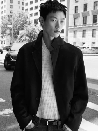 East Asian male model posing on a city street wearing a black coat over a white textured knit sweater for COS Fall 2026.