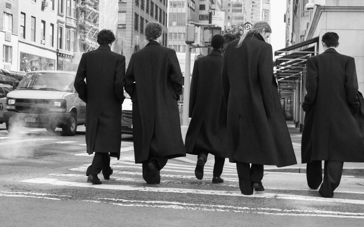 Five models seen from behind in matching long black tailored overcoats walking away across an urban crosswalk in black and white.