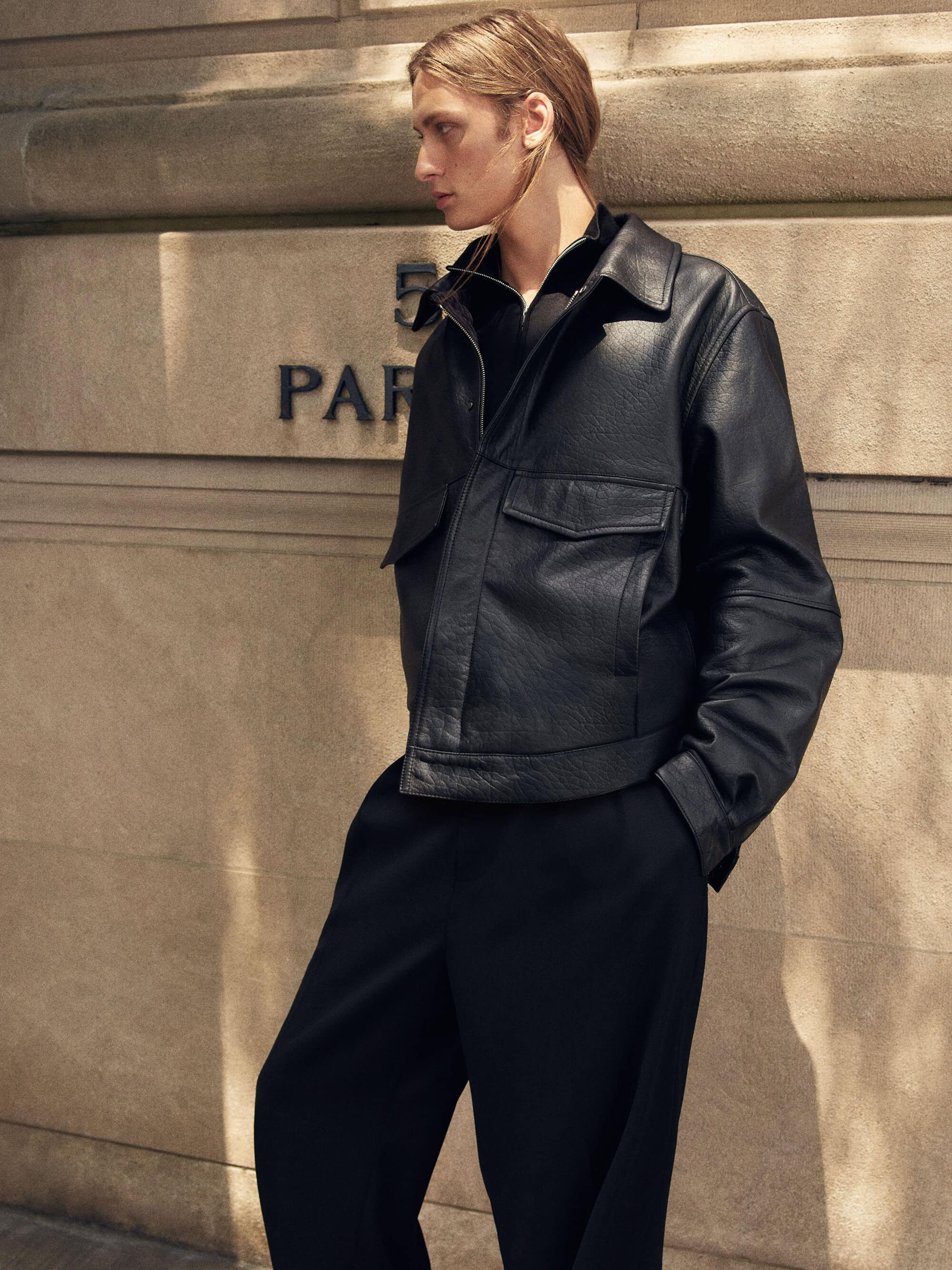 Model standing outdoors against a stone building wall wearing a structured black leather jacket and black trousers.