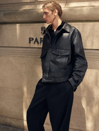 Model standing outdoors against a stone building wall wearing a structured black leather jacket and black trousers.