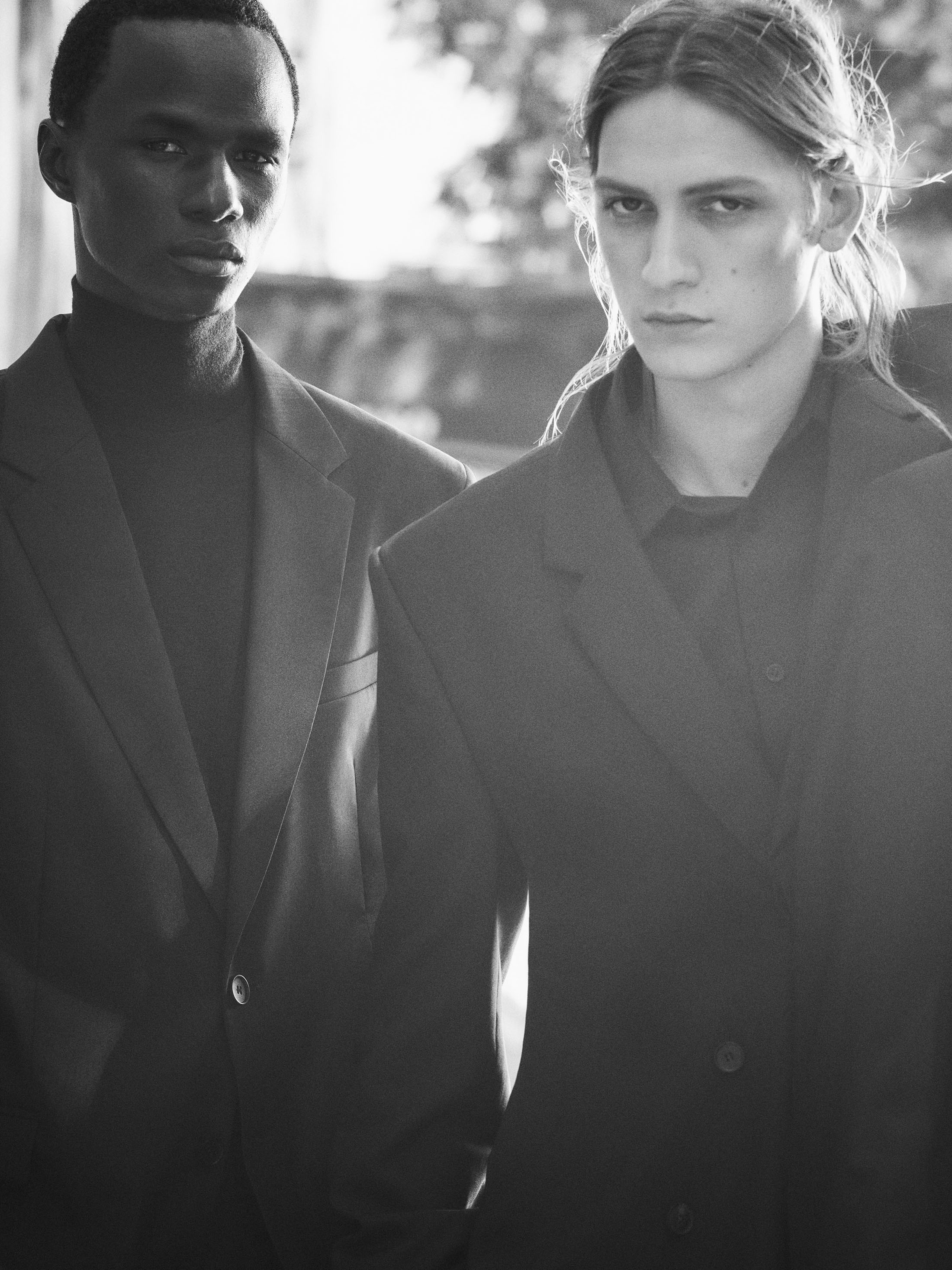Black and white portrait of two male models wearing dark tailored blazers and high collars under natural backlight.