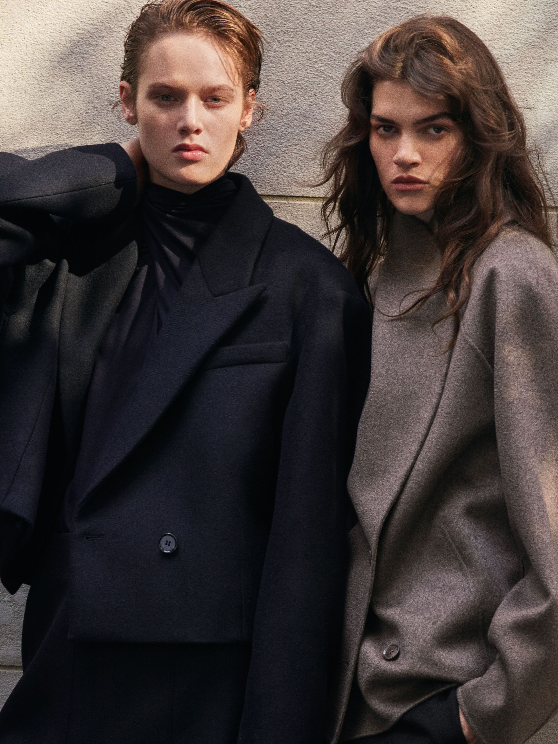 Two models posing outdoors in structured black and taupe wool jackets for COS Fall 2026 campaign against a textured neutral wall.