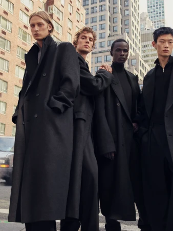 Four male models wearing tailored black overcoats standing on a city street surrounded by tall buildings.