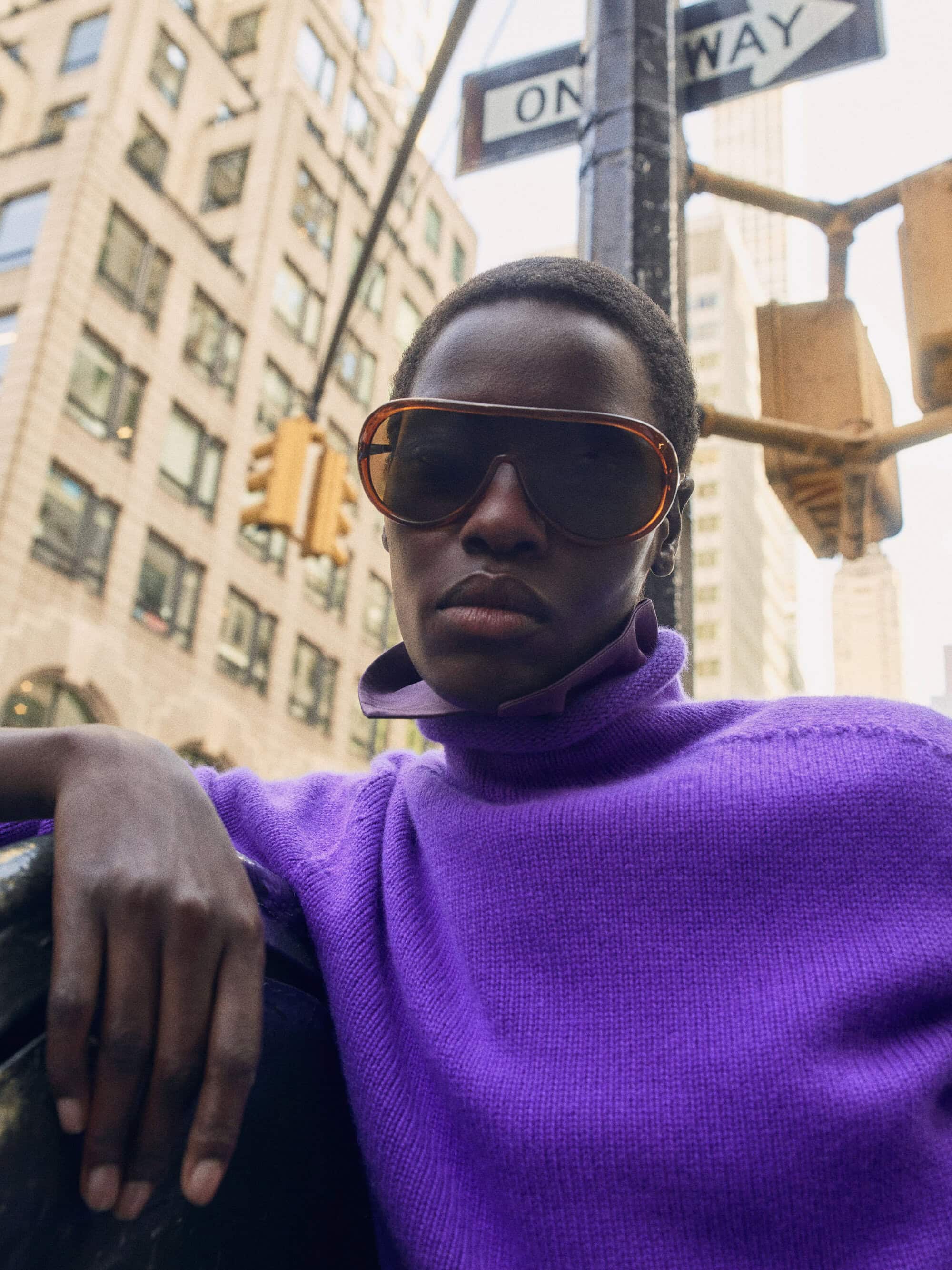A person with short hair wearing large tinted sunglasses and a vibrant purple turtleneck sweater outdoors in a city setting.