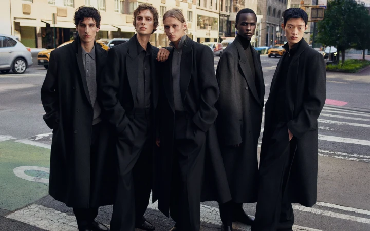 Five male models wearing long black tailored overcoats and trousers stand together across an urban city crosswalk.