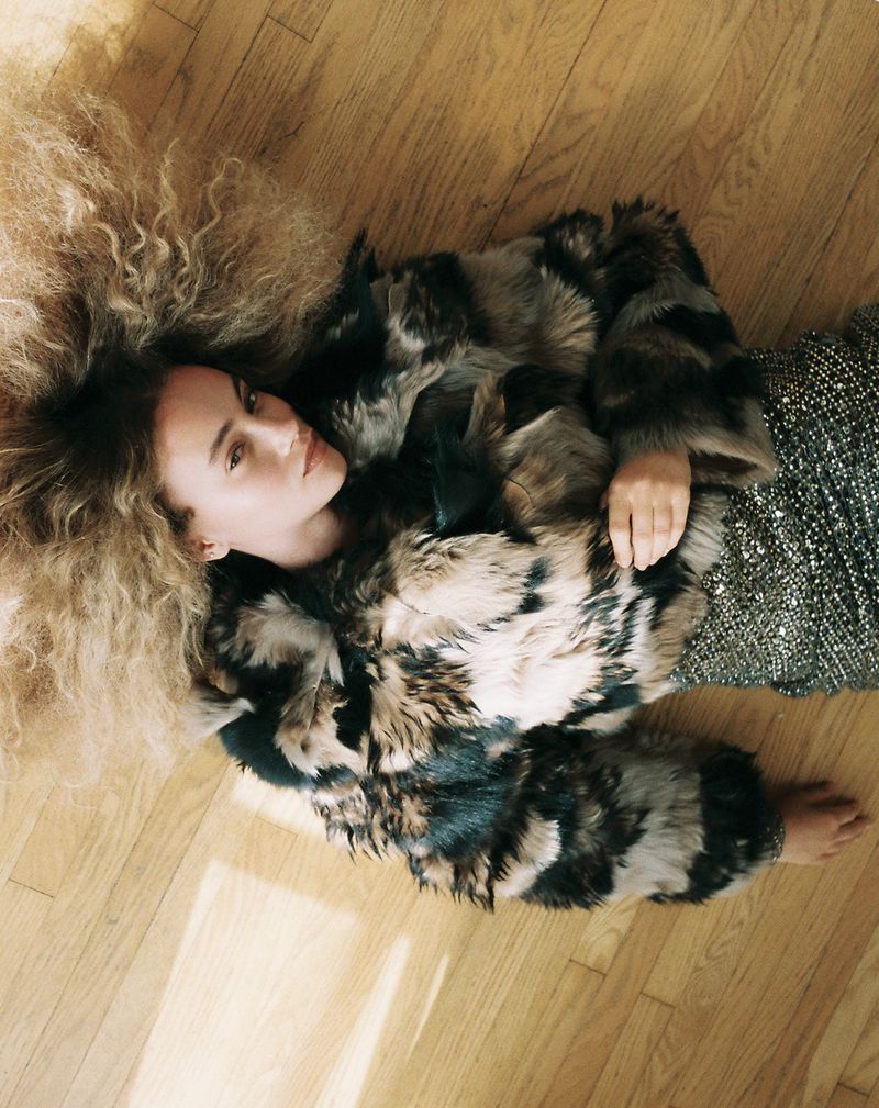 Woman with voluminous crimped hair lying on a wooden floor in a plush patterned fur jacket and sequined skirt.
